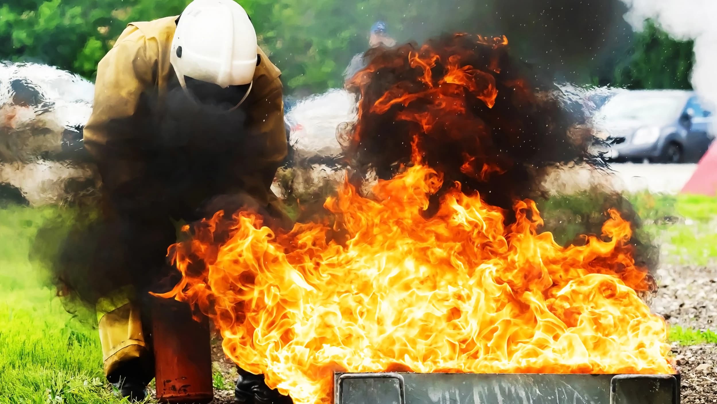 Feuerwehrmann bei Übung, lodernde Flammen, dichter Rauch, Schutzausrüstung, heller Schutzhelm, dunkle Jacke, dynamische Szene.